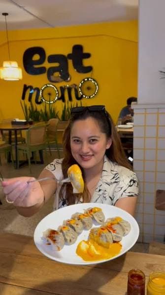 A guest holding up a plate of jhol momo in front of the yellow eat momo wall